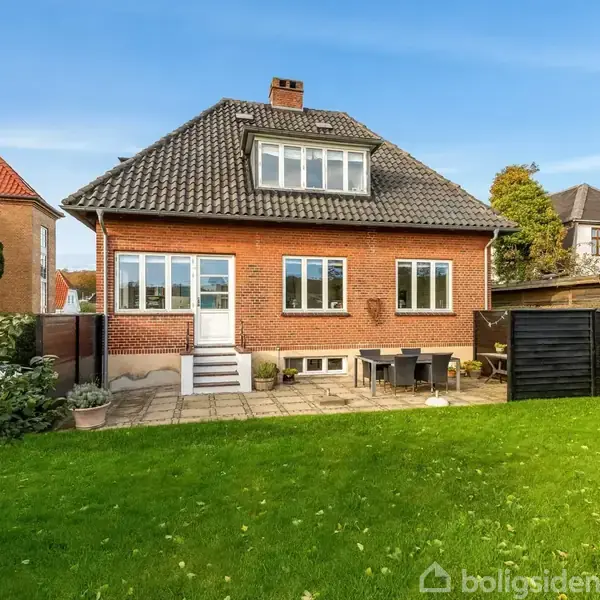 Et rødt murstenshus med mørkt tegltag og kvist står i en grøn have. Foran huset befinder sig en fliseterrasse med havemøbler.