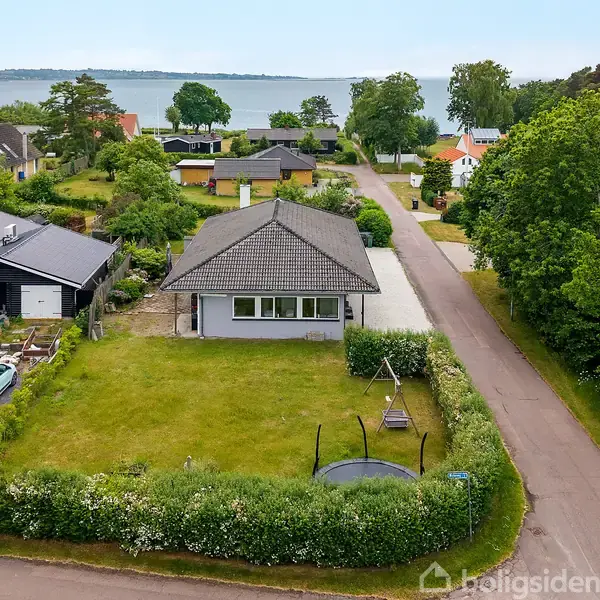 Et enkelt hus står på en grøn grund med en trampolin i haven, omgivet af en stille vej og naboens huse. I baggrunden ses en sø og træer.