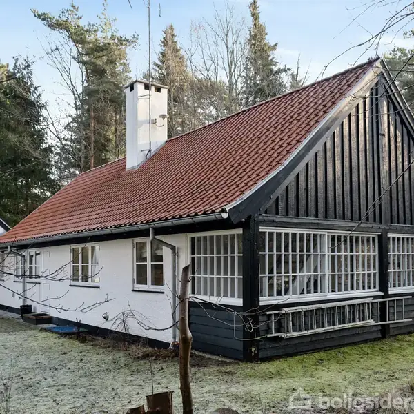 Hus med rødt tegltag og hvide vægge står stille omgivet af nåletræer. Store vinduespartier præger facaden, og en skorsten ses på taget. Frostbeklædt græsplæne i forgrunden.