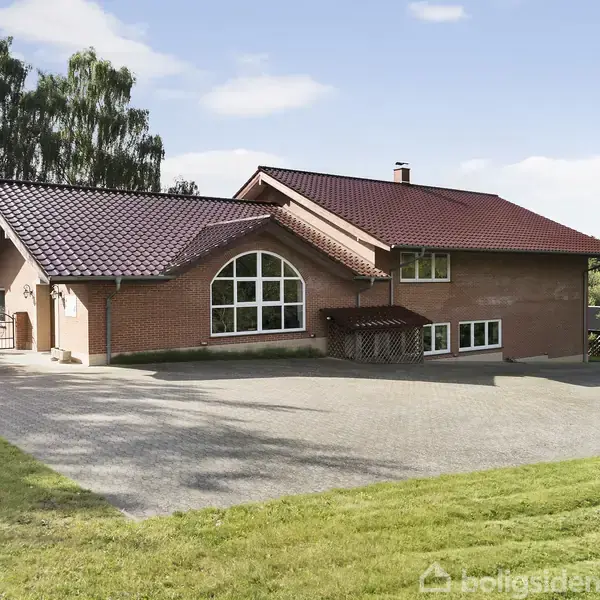 Et rødt murstenhus med skråt tag og stor buevindue står på en stenbelagt indkørsel omgivet af grønne græsarealer og træer under en blå himmel.