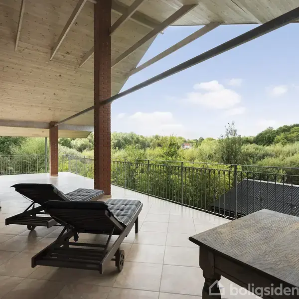 To liggestole står på en flisebelagt terrasse under et træloft, med udsigt til frodig grøn bevoksning og en lys blå himmel i baggrunden.