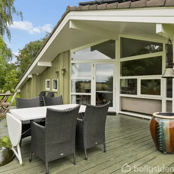 Et udendørs terrasseområde med et bord og fire sorte stole på en træveranda. Store glasvinduer leder indenfor i et hus omgivet af grønne planter og træer.