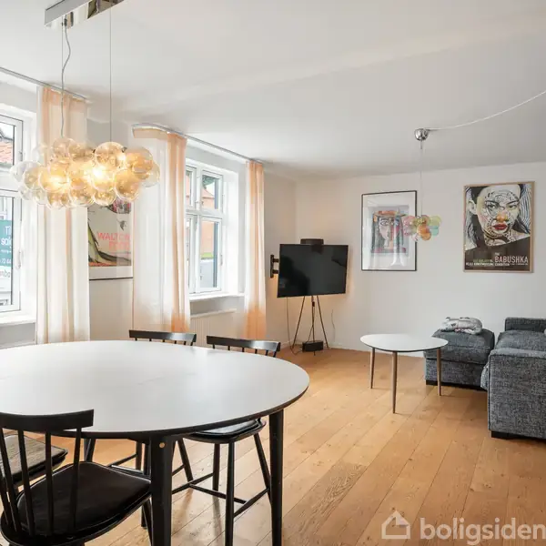 Rundt spisebord med sorte stole står i en lys stue med trægulv. Sofa ved siden af et sofabord og et TV på stativ. Malerier hænger på væggen.