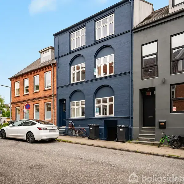En række byhuse står langs en stille gade. Et blåt treetagers hus med buede vinduer ses. En hvid bil er parkeret på vejbanen. Cykler står op ad husene.
