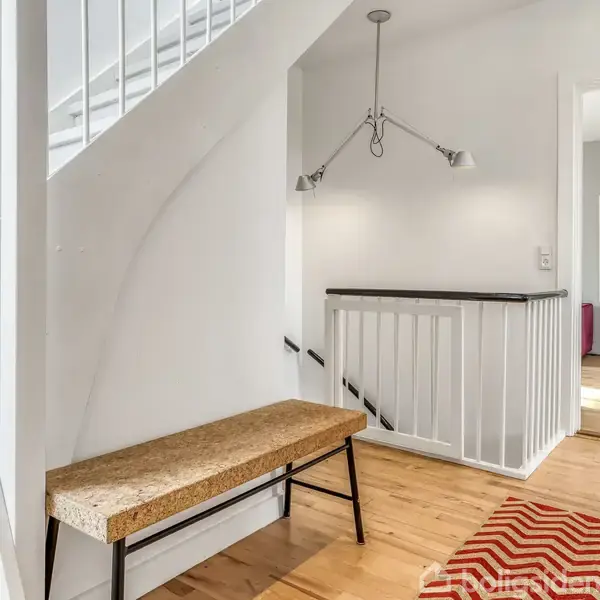 Bænk af træ står i en lys gang ved siden af en trappe med hvidt gelænder. Lysebrunt trægulv og vægmonteret lampe skaber en minimalistisk indretning.