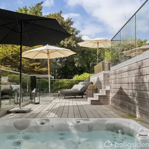 Boblebad udenfor med vand, omgivet af terrasse af træ og glasrækværk. To parasoller skaber skygge over liggestole. Trin fører til en hævet del med planter og mere siddeplads.