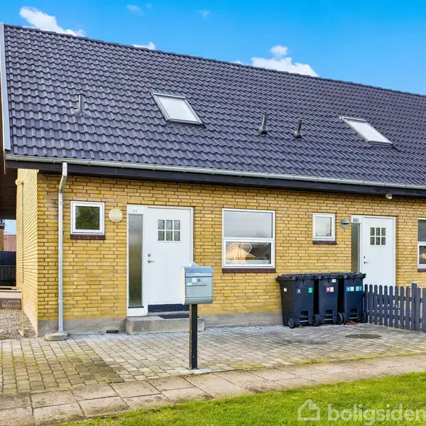 Et rækkehus med gul murstensfacade står i en boligkvarter. Flere skraldespande står foran huset. Tætklippede hække omgiver ejendommen og en klar blå himmel ses i baggrunden.