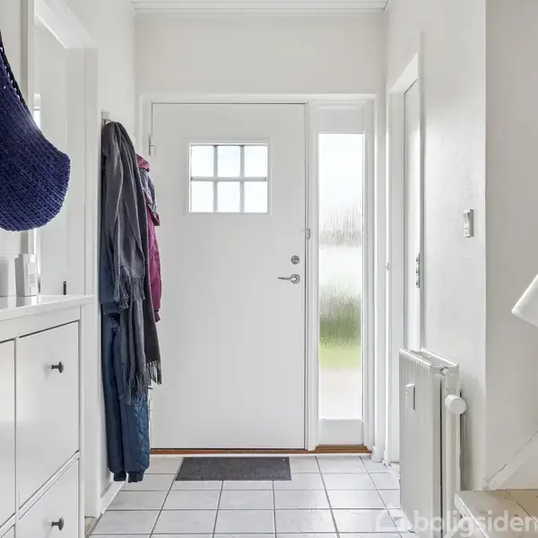 Dør i entré åbner ud mod hvidmalede vægge og flisegulv. En skohylde står til venstre, med jakker hængende ved siden af. En blå kurv og lille plante ses ovenpå hylden.