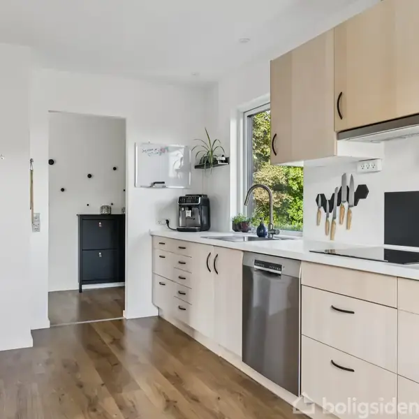 Et lyst køkken har lyse træskabe og en opvaskemaskine. En kaffemaskine står ved siden af en vask under et vindue. En åben dør fører til et andet rum med mørke møbler.