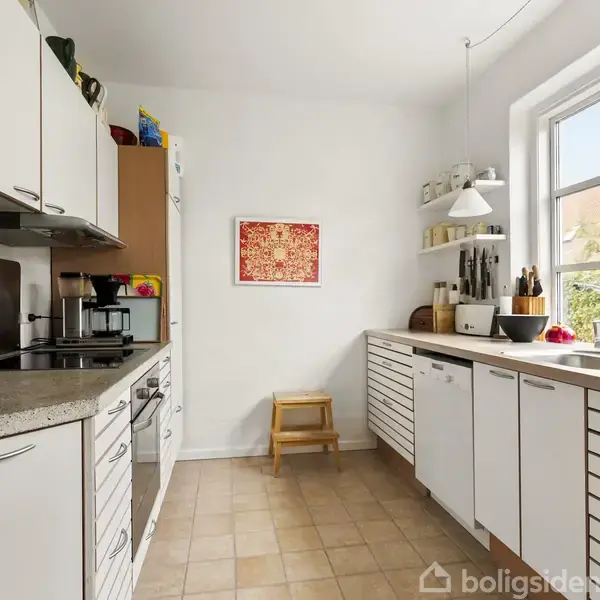 Et moderne køkken med hvide skabe og bordplader i træ. Kaffemaskine og knivblok står på bordet. En rød dekoration hænger på væggen over en træskammel.