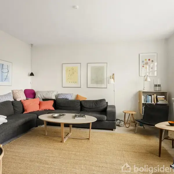 En stue med en stor, mørkegrå hjørnesofa fyldt med farverige puder. Et rundt sofabord foran sofaen. Væggene har kunstværker, og en bogreol står ved siden af vinduet.