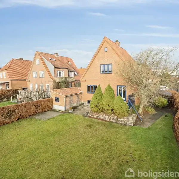 Et rødt murstenshus står stille omkranset af en velplejet græsplæne og hække. Omkringliggende huse ses i baggrunden under en klar blå himmel.