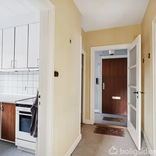 Et køkken med hvide skabe og en ovn står til venstre. En gang fører til en brun hoveddør i en entré med tæppe.