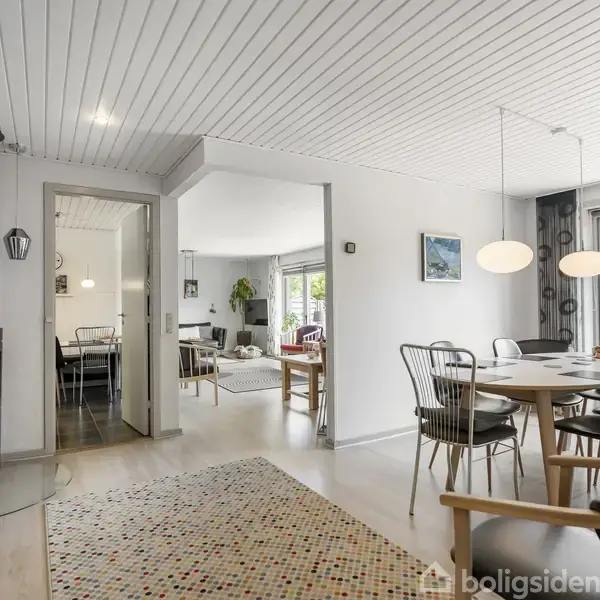Brændeovn står tændt i hjørnet. På højre side er der et spisebord med stole og to hængelamper. Store vinduer lyser det moderne opholdsrum med hvidt loft op.