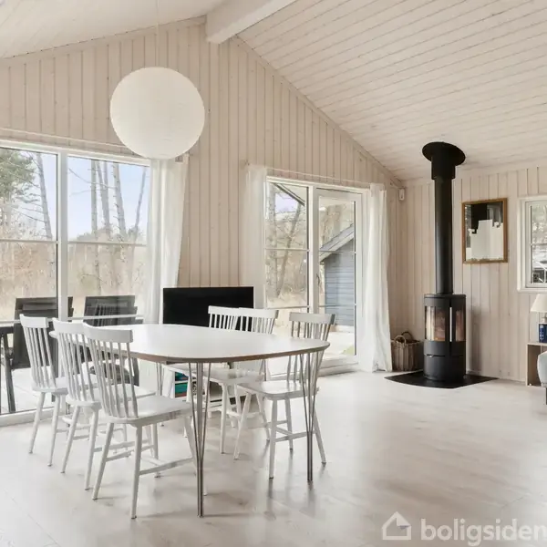 Et lyst og moderne rum med et spisebord og hvide stole foran store vinduer. En pejs står i hjørnet ved siden af et gråt sofahjørne, omgivet af træpanelvægge.