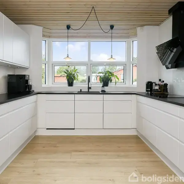 Hvidt, moderne køkken med sorte bordplader; to planter står i vindueskarmen over vasken. Træloft, trælaminatgulv og hængelamper skaber en lys, ren atmosfære. Mixere og kaffemaskine til venstre.