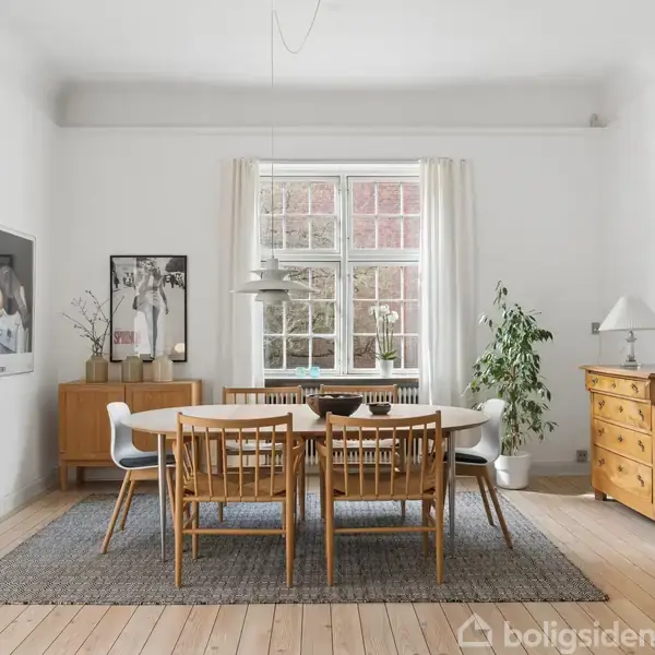 Et spisebord står midt i et lyst rum med træstole omkring. Et tæppe ligger under bordet. Væggene er pyntet med kunst. Planter og en kommode kompletterer indretningen.