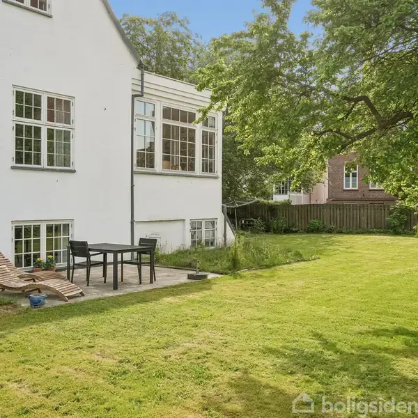 Hvidt hus med vinduer, står stille i en have med grønne græsplæner. Havemøbler er placeret tæt på bygningen, omgivet af træer og en anden bygning i baggrunden.