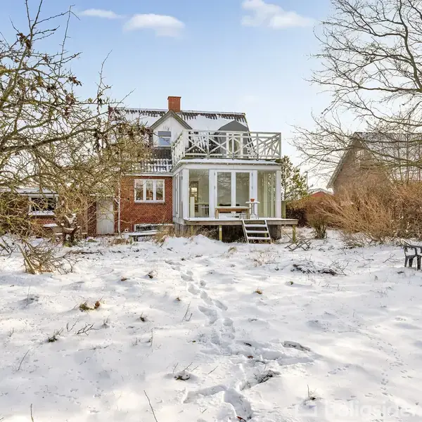 Et rødt murstenshus står i en snedækket have med spor i sneen. Et træ med bare grene og et bord med paraply ses i forgrunden.