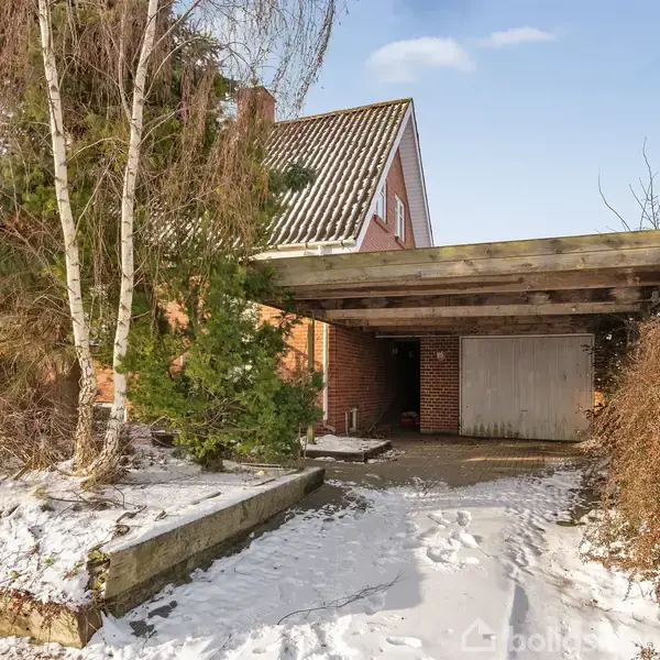 Et rødt murstenshus med garage i sneklædt landskab. Garagetaget er fladt og omgivet af buske og træer. Skinnede, klar vinterhimmel over. Ingen tekst fundet i billedet.