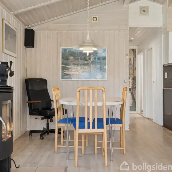 En brændeovn står i et lyst køkkenalrum. Ved siden af står et rundt spisebord med fire stole. En sort kontorstol er placeret til venstre, og et maleri hænger på væggen.