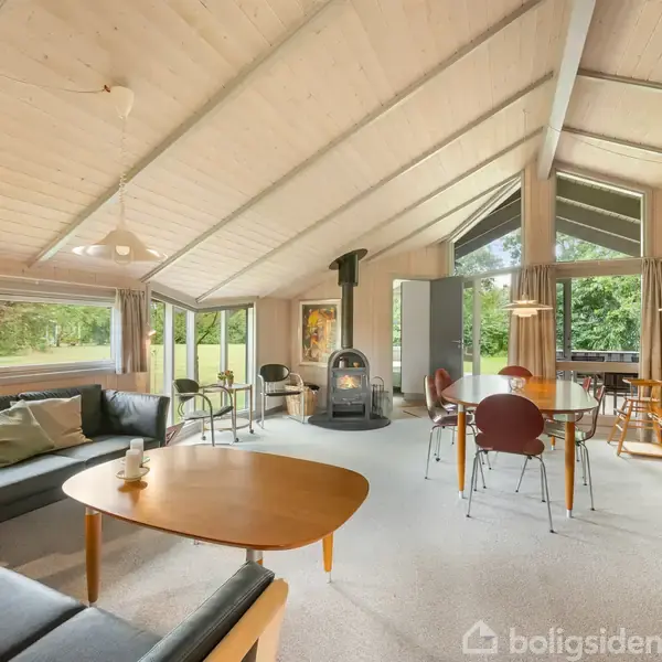 Stue med moderne møblering; sorte sofaer, træbord og brændeovn i midten. Store vinduer slipper lys ind, mens græsarealet er synligt udenfor. Pejse-området understreger hygge og enkelthed.