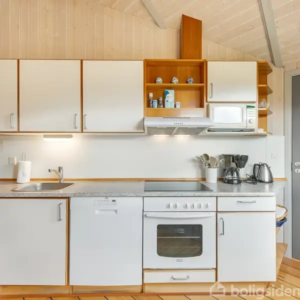 Et køkken med komfur og ovn indbygget i hvid køkkenenhed, omgivet af skabslåger og åbne hylder. Der er diverse køkkenapparater som mikroovn, kaffemaskine og elkedel. I baggrunden ses en stue med spisebord.