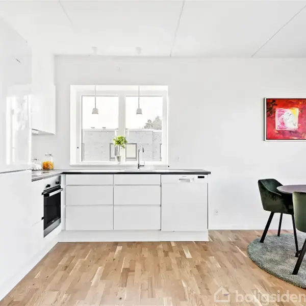 Et stilrent, lyst køkken med hvid køkkenindretning, træbordplader og spiseplads med runde bord, grønne stole. Store vinduer giver naturligt lys, og enkel kunst pryder væggen. Moderne interiørdesign.