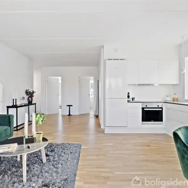 Stue med grøn sofa og sofabord, står ved siden af åbent køkken i hvidt. Rummeligt med trægulv, store vinduer og hvide vægge, tilbyder moderne og lys atmosfære.