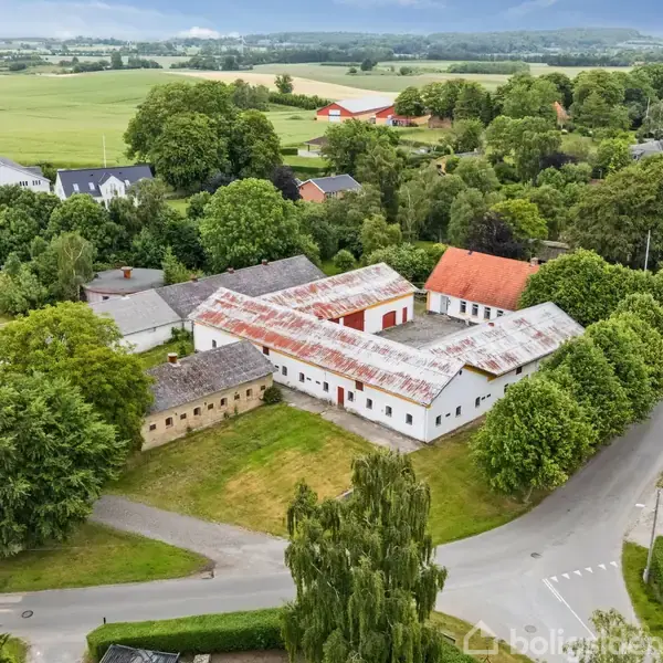Hvidt hus med rødt tegltag, omgivet af frodige buske og et græsområde. Huset har fem vinduer og ligger i en have med træer i baggrunden.