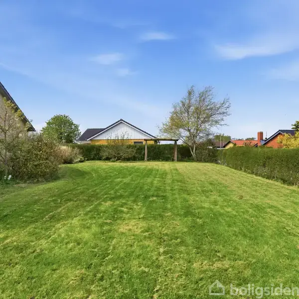 En grøn græsplæne strækker sig mellem hække, med et hus til venstre og trækonstruktioner i baggrunden under en blå himmel.