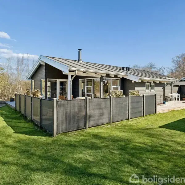 Et moderne, gråt hus med store vinduer står omgivet af en grøn have. Huset har en indhegnet terrasse og flere blomsterkasser. Træer og en skov er synlige i baggrunden.
