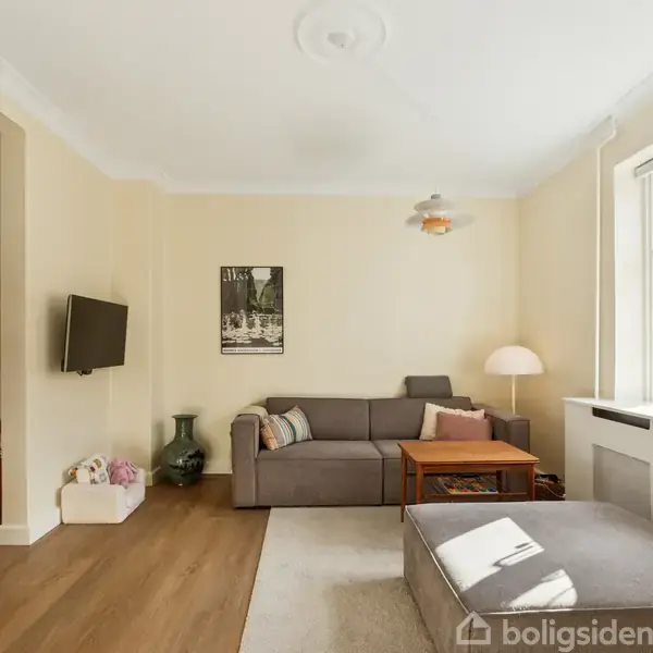 Sofa står i et lyst stuehjørne ved et stort vindue, omkranset af lamper og dekorationer. TV er vægmonteret til venstre, og et tæppe ligger delvist på trægulvet.