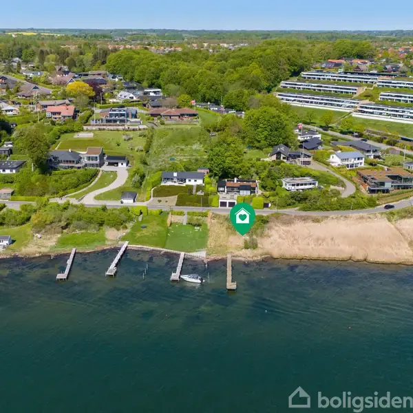 Et kystnært område med huse og bådebroer, omgivet af frodig vegetation og grønne arealer. Bygninger ligger tæt ved vandet, og der er en blanding af beboelseshuse og større, mere moderne strukturer.