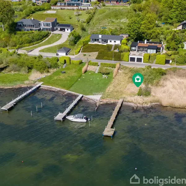 Kystnært område med tre bådebroer strækker sig ud i vandet. Små huse med haver er beliggende på bakken over for vandet, omgivet af grønne træer og buske.