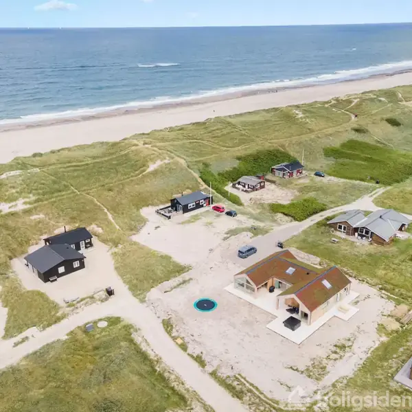 Flere sommerhuse står stille i et klitlandskab nær kysten med havet i baggrunden. Spredt vegetation omgiver husene, og stier forbinder dem til stranden.