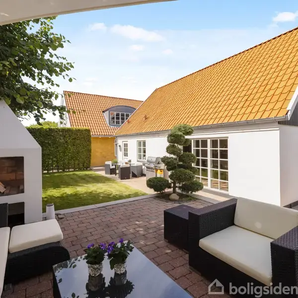 Terrassemøbler står på en flisebelagt patio foran et hvidt hus med orangetræstag. I baggrunden ses en udendørs pejs, græsplæne og velplejede buske under en blå himmel.