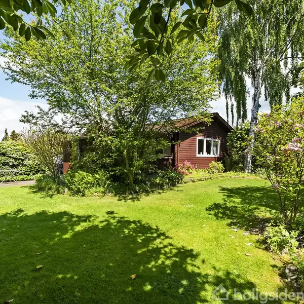 Et rødt træhus står delvist dækket af frodige træer og buske i en velholdt grøn have under en klar blå himmel.