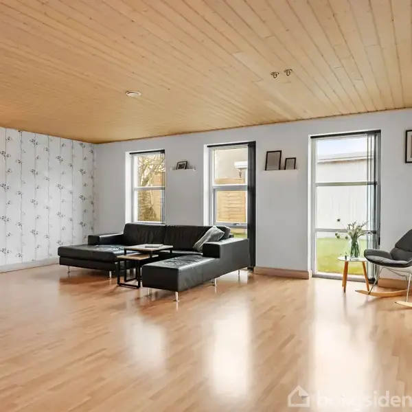 Sort sofa står i lys stue med træloft, parketgulv og tre store vinduer. Rum dekoreret med mønstrede tapeter og moderne stole.