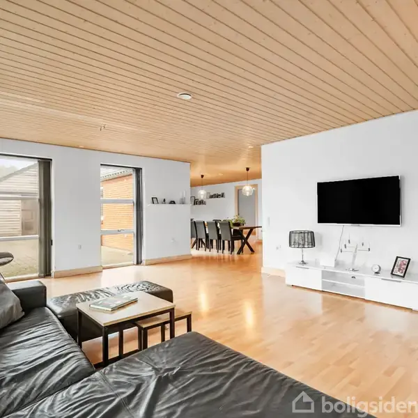 Sort lædersofa står i en rummelig stue med lyst trægulv og træloft. Her er et vægmonteret TV, lavt hvidt tv-møbel og store vinduer med udsigt til en terrasse.