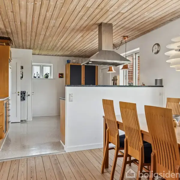 Køkken-alrum med træloft og gulv. Et bord med stole står ved siden af en halvåben køkkenø med emhætte. Hylder med bøger og planter er på væggen. En moderne lampe hænger over bordet.