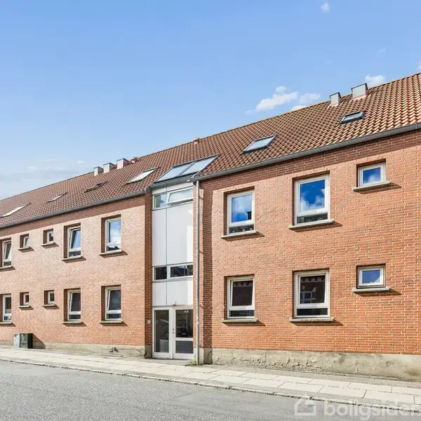 Rød murstensbygning med brune tegltag, vinduer i gavlen og facade, står på en stille gade. Det klare vejr kaster lys over bygningen og den asfaltbelagte vej.