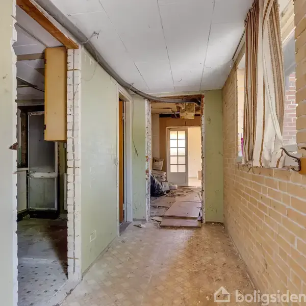 Gang med murstensvægge og nedhængt loft. Gulvet er dækket af plastik eller laminat. Vinduer med mønstret gardin til højre. Døråbning forrest fører til lysere rum.