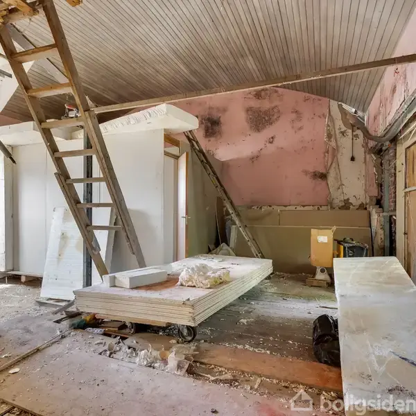 Et faldefærdigt rum med byggeplader og stige mod en væg. Maling skaller af de lyserøde og blå vægge. Byggerod fylder gulvet, hvilket indikerer renovering.