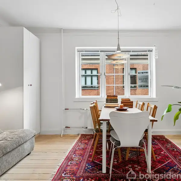 Et spisebord med stole er placeret på et rødt tæppe. Bordet er dækket og har en pendel over sig. Til højre står en grøn plante ved siden af et vindue.