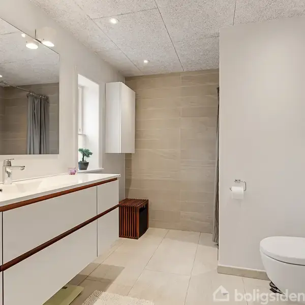 Moderne badeværelse med stor håndvask og skab langs venstre væg. Toilet til højre, vægmonteret med toiletpapirholder. Neutrale fliser, loftsbelysning og et vindue giver lys.