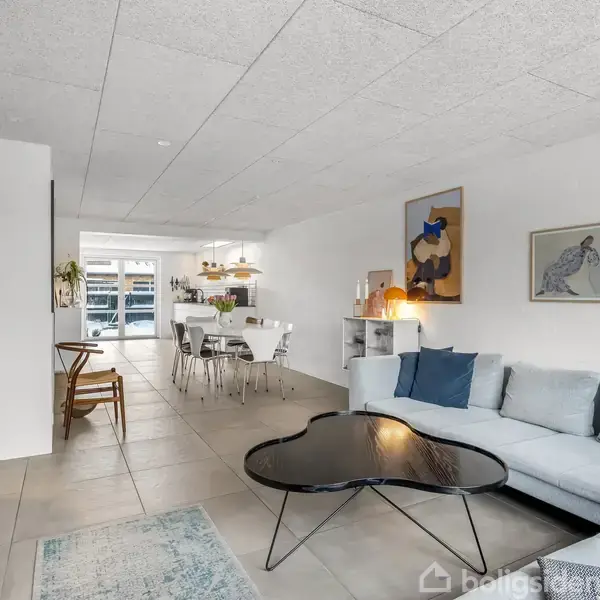 Stue med en grå sofa og et sort sofabord. Et spisebord med hvide stole står i baggrunden. Væggene prydes af moderne kunstværker i et lyst, åbent rum.