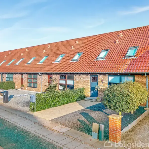 Rækkehus står langs en gade med rødt tegltag og flere vinduer i taget. Foran er der en lille have med buske, omgivet af en gangsti og parkeringsområde.