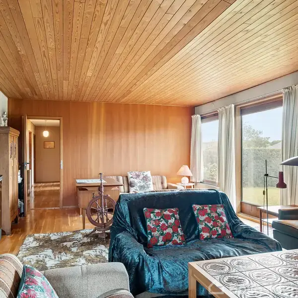 Stue med sofa og lænestole placeret nær et stort vindue med udsigt. Træloft og -vægge skaber en hyggelig atmosfære. Et tv er placeret til venstre, og et rosenmønstret tæppe dækker gulvet.