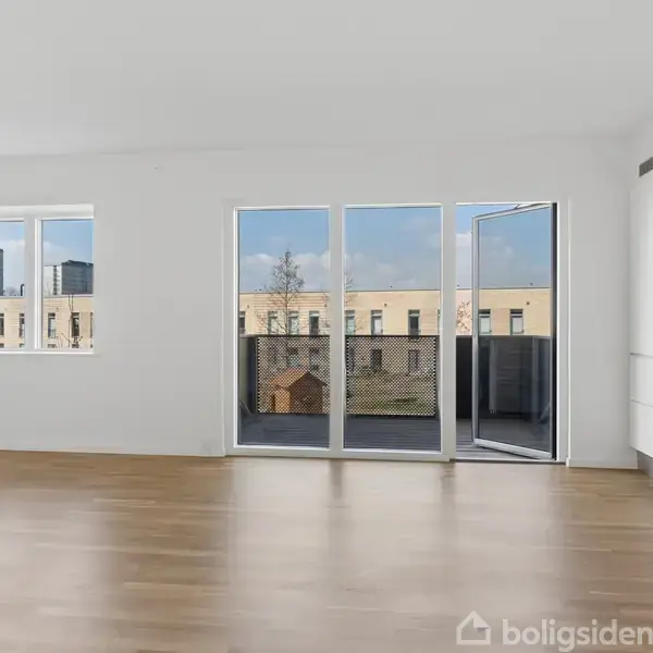 Rummelig stue med lyse vægge og trægulv åbner op til en balkon gennem en glasdør. Store vinduer lukker naturligt lys ind, og et moderne, hvidt køkken er på højre side.